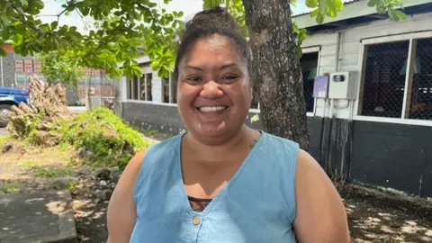 Samoan Teacher Amanda Taavaomaalii. She is wearing a blue vest and smiling into the camera while standing in a garden by a single-storey building.