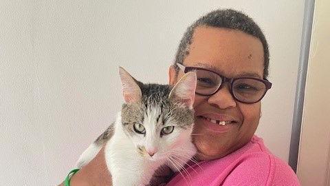 Patsy Wright with short dark hair and glasses in a pink T-shirt, holding a tabby and white cat. She is smiling at the camera.