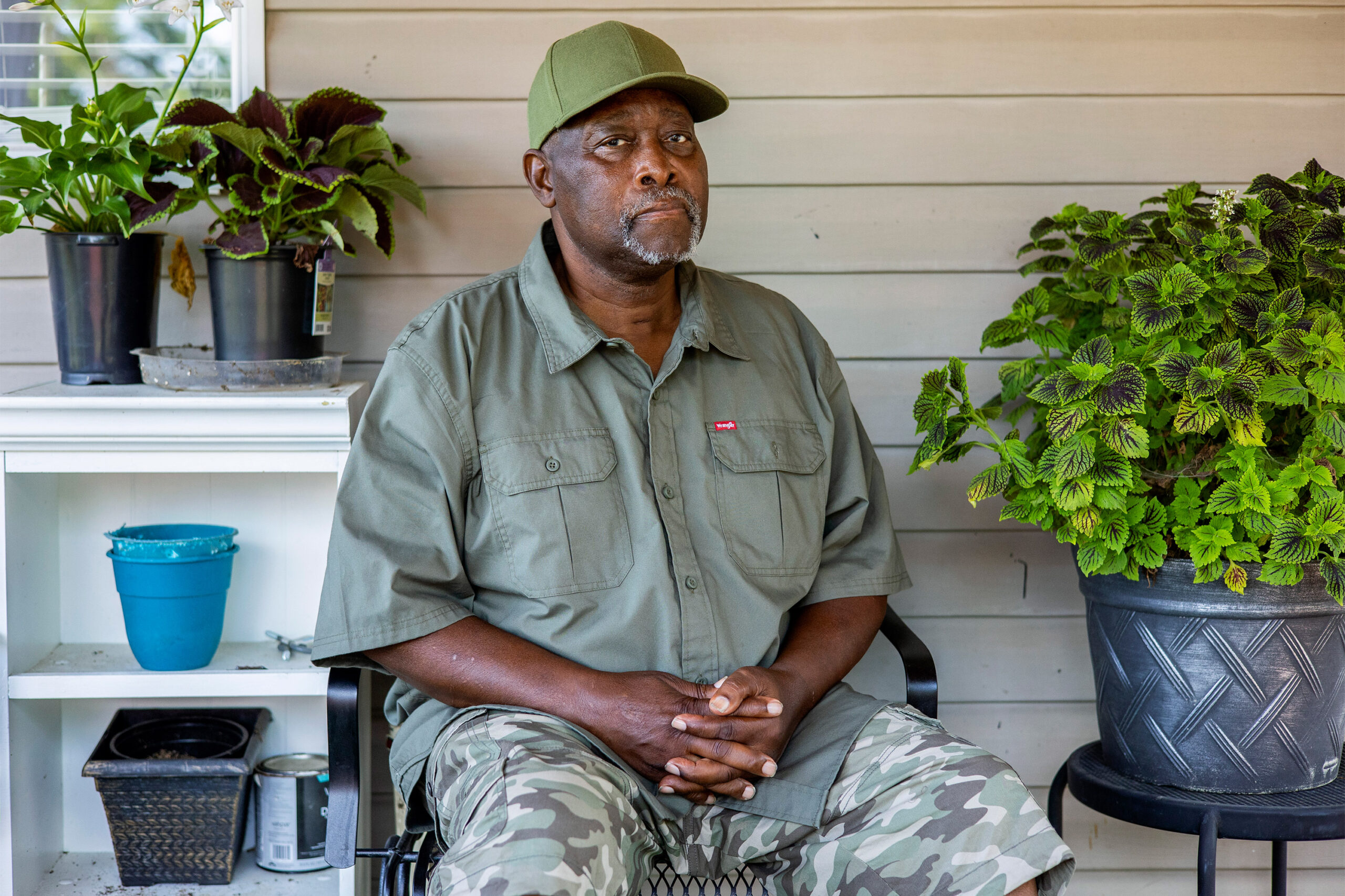 A photo of a Black man sitting on a porch. To his left and right are potted plants.