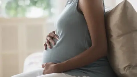 Getty Images A pregnant woman sitting down cradling her bump. She is wearing a grey, sleeveless top and you cannot see her head.