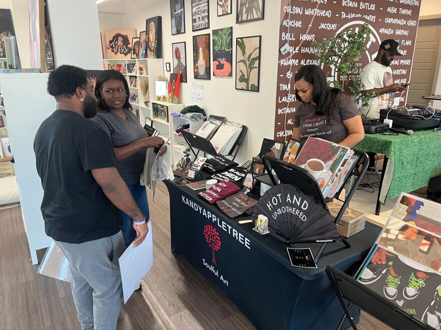 A woman on the right has a table covered in merchandise as two people on the other side of the table speak to each other.