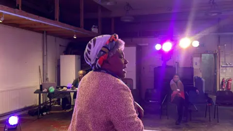 The character of Miss Ida in rehearsals on stage. She is wearing a hat and is being watched by two people sitting down as lights project onto the stage area.