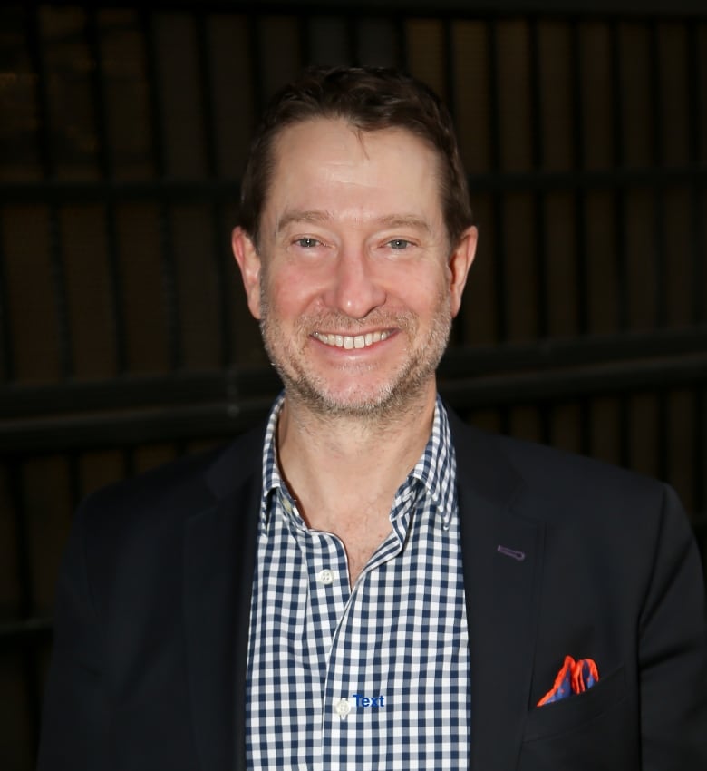 A man with dark hair and short facial stuble wears a dark blazer and check pattern shirt, smiling at the camera. 