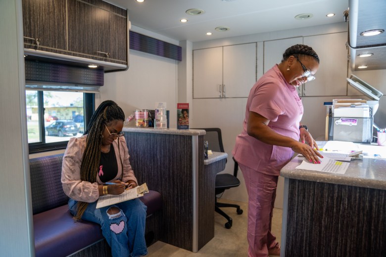 A seated Black woman fills out paperwork on a clipboard while another Black woman in pink scrubs looks over paperwork on a countertop. 