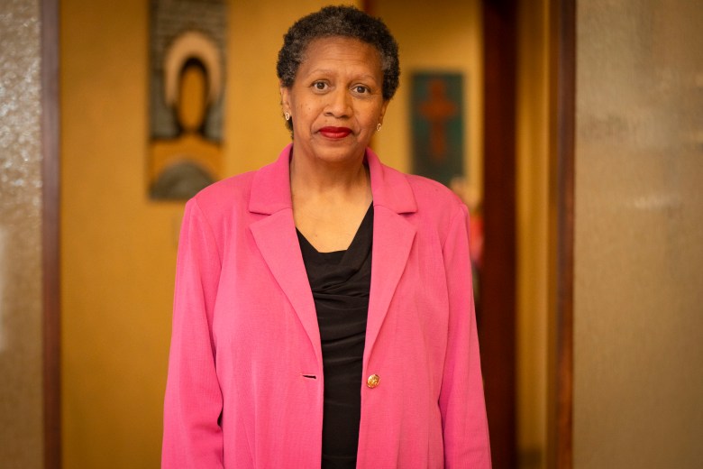 A Black woman in a black shirt and pink suit jacket stands for a portrait.