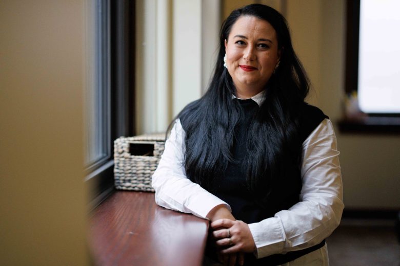 A woman with long dark hair, wearing a black vest over a white shirt, stands by a window ledge, looking at the camera. A woven basket is on the ledge beside them.