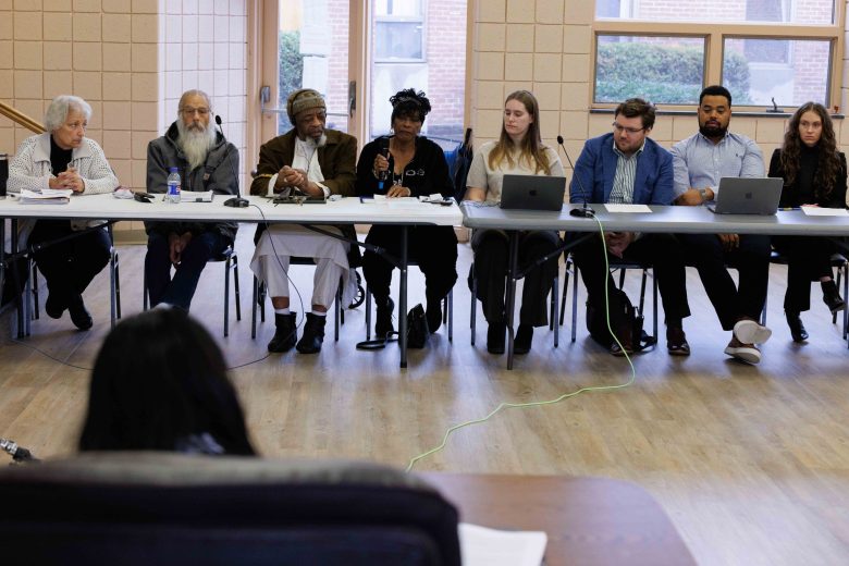 A group of people sitting at a long table with laptops and microphones, engaging in a discussion in a community meeting room.