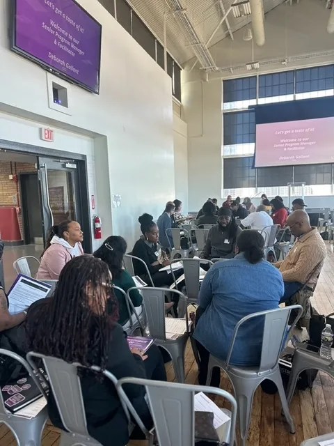 A group of people seated in a large room, participating in a workshop or seminar, with a presentation slide visible on a screen.