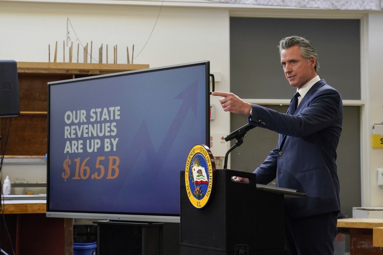 A person wearing a navy blue suit points in front of them as they stand behind a lectern with the California seal in front of it. A large screen with a graphic that reads 