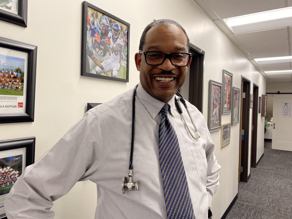 A Black man in glasses with a stethoscope draped over his shoulders smiles at the camera. He is wearing a blue, diagonally striped tie and standing in a hallway lined with pictures of the Denver Broncos