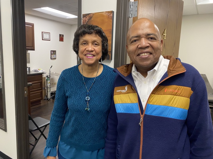 A Black woman with short wavy hair in a blue knit sweater stands on the left of a bald Black man wearing a blue fleece jacket over a white dress shirt. Both are looking at the camera and smiling. There are standing in the doorway of a patient room in a doctors office.