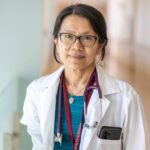 A female physician wears a lab coat in a hospital hallway. She has a stethoscope and a lanyard around her neck and a cell phone in her pocket.