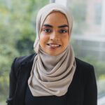 Headshot of Fatima Sheikh. She wears a black sweater and a beige hijab, standing in a hospital hallway.