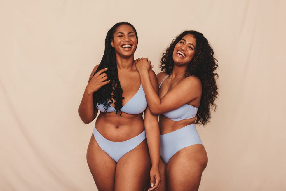 friends celebrating their natural bodies in a studio. two happy young women laughing cheerfully while wearing blue underwear. body positive young women standing together against a studio background.